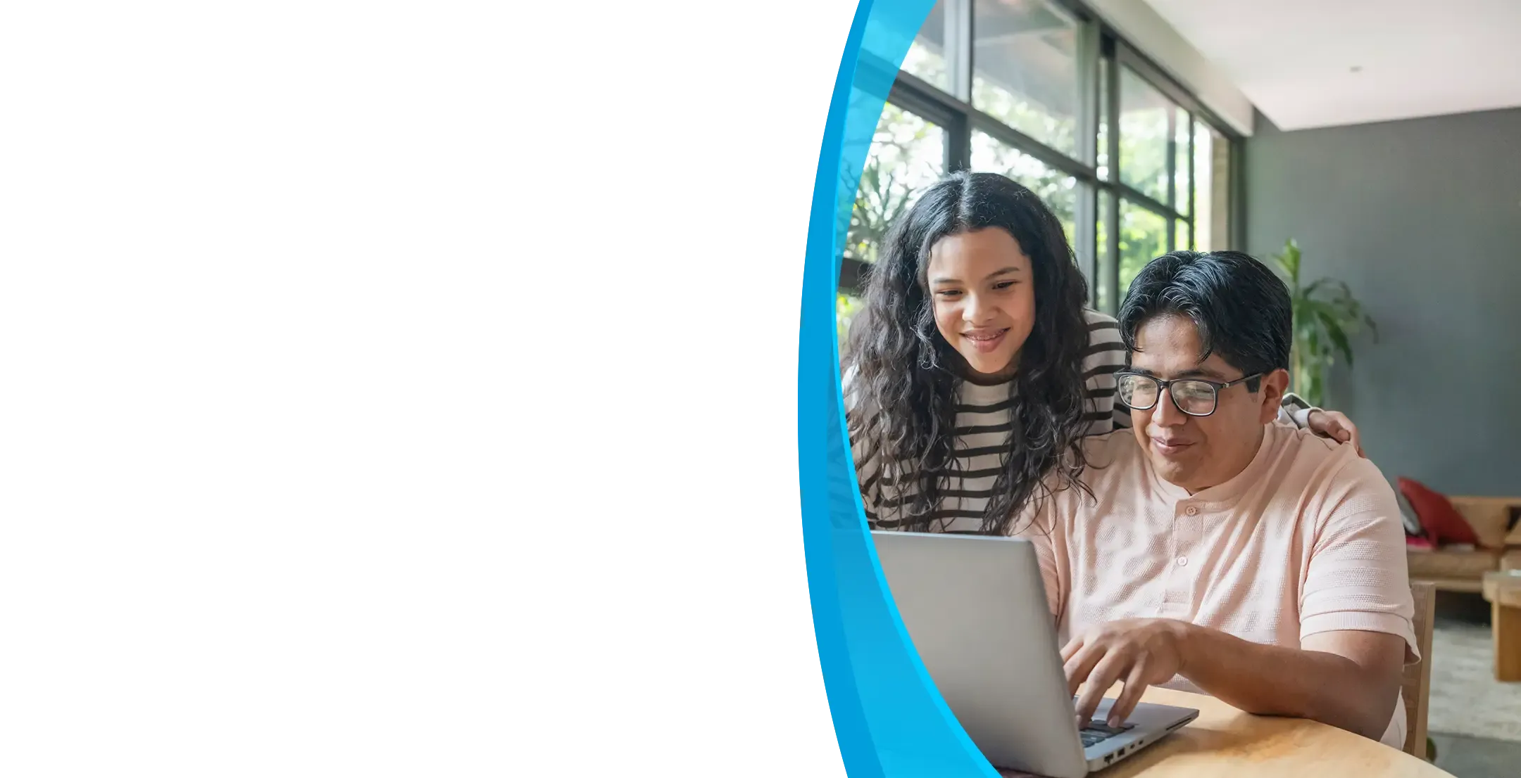A man and teenage girl smile together while using a laptop at a home dining table.